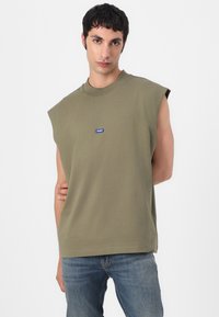 Camiseta sin mangas verde oliva, de material de algodón, con cuello redondo, que presenta un pequeño logotipo azul "HUGO" en el frontal. Diseño sencillo y de corte relajado.