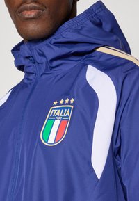 Námornícka modrá vietorodolná bunda s kapucňou, so zlatým zipsom a bielymi detailmi. Na prednej strane sa nachádza logo talianskej národnej futbalovej reprezentácie s štyrmi hviezdami nad ním.