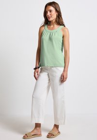 Zelený top bez rukávů se shromážděním, kombinovaný s volnými bílými kalhotami a hnědými tkanými sandály. Hladká látka, uvolněný střih.