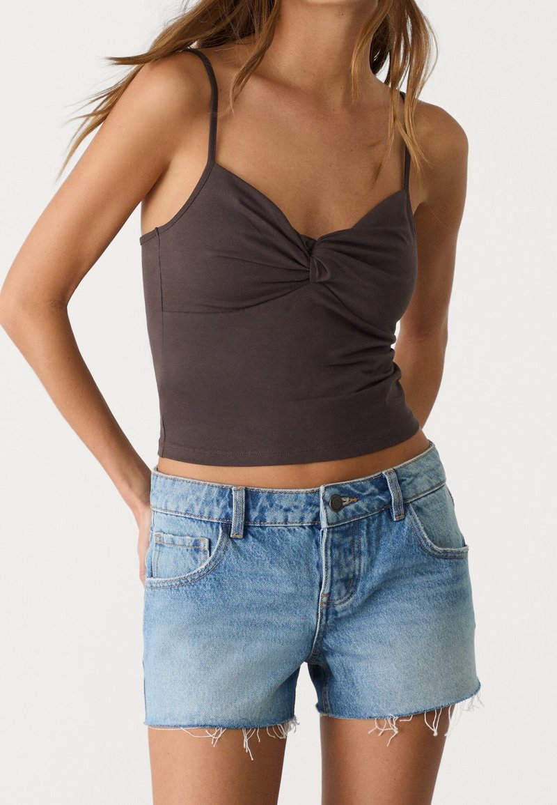 Camiseta sin mangas ajustada de color gris oscuro con un detalle frontal retorcido, combinada con shorts de mezclilla azul claro con dobladillo deshilachado y bolsillos clásicos.