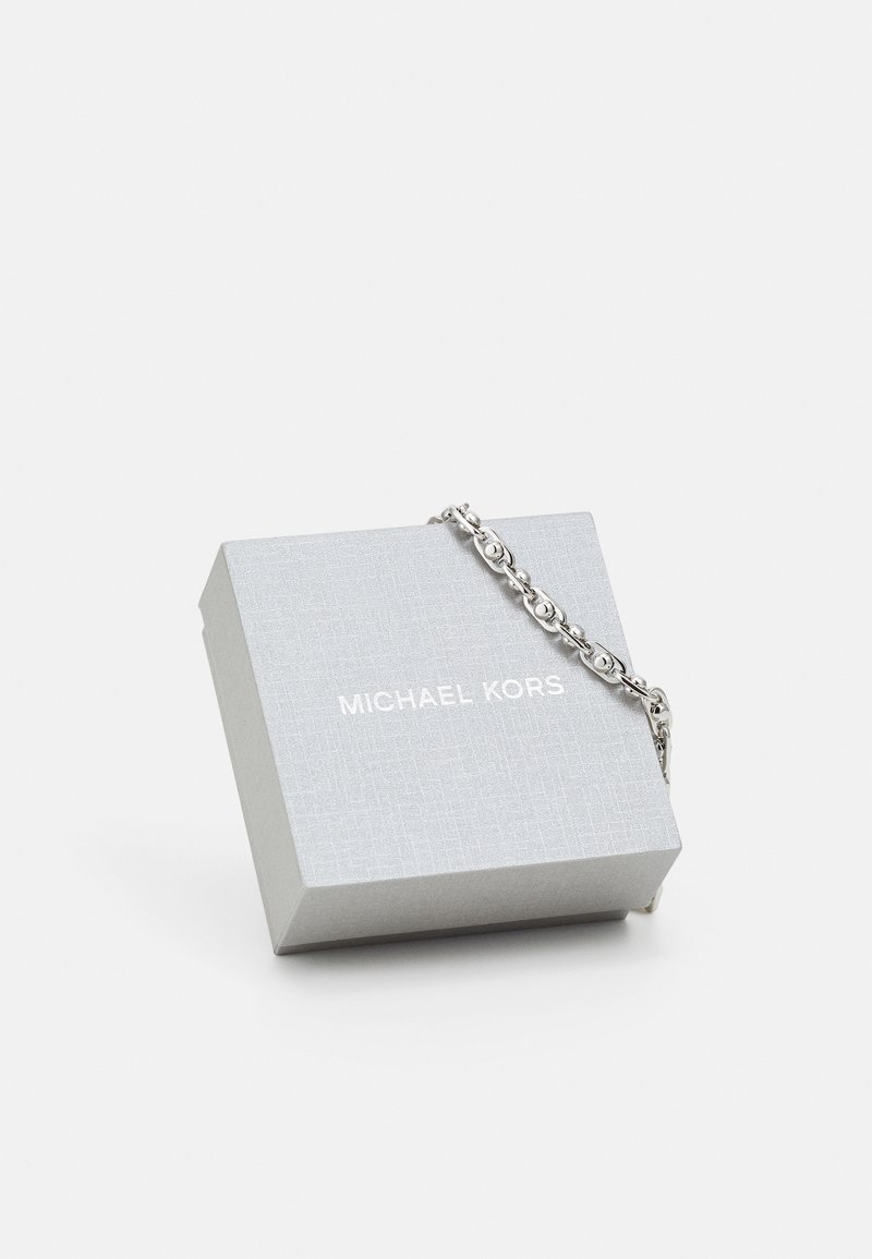 Zilveren armband die een gestructureerde, lichtgrijze geschenkdoos omcirkelt met het "Michael Kors"-logo in het wit op de bovenkant gedrukt.