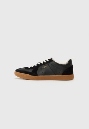Black leather and suede sneaker with a gum outsole, white laces, and embossed logo detail on the side. Features a low-profile design.