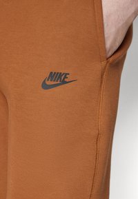 Nike trenirke u toploj smeđoj boji, s glatkom teksturom i diskretnim crnim logom s lijeve strane.