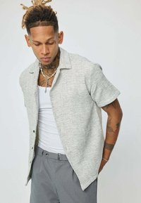 Chemise à manches courtes, gris clair et texturé, avec un col pointu et une fermeture à boutons sur le devant, portée sur un débardeur blanc et un pantalon gris.