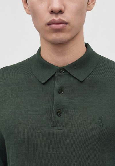 Gros plan d'une personne portant un polo en maille vert foncé avec trois boutons et un petit logo brodé sur la poitrine.