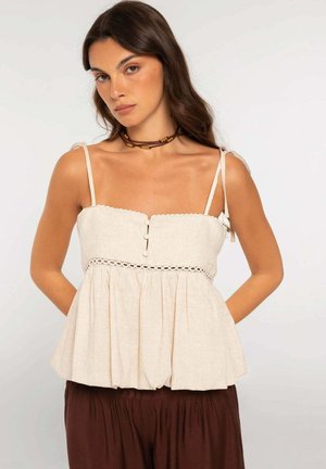 Mujer con cabello largo y castaño que lleva una camiseta sin mangas beige con botones y detalles de encaje, combinada con pantalones sueltos de color marrón oscuro.