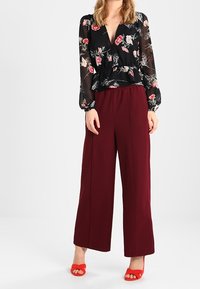 Donna che indossa una blusa nera a maniche lunghe con motivo floreale, pantaloni bordeaux a vita alta e ampia gamba, e scarpe con tacco rosse a piedi scoperti, in piedi contro uno sfondo bianco.