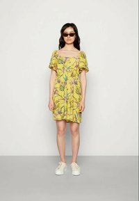 Vestido floral amarelo com mangas curtas, cintura franzida e comprimento acima do joelho. Combinado com ténis brancos e óculos de sol escuros.