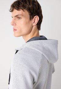 Un sweat à capuche blanc avec un motif de points noirs, présentant une capuche à bord gris et une fermeture éclair, exposé de côté.
