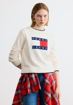 Fiatal nő fehér Tommy Jeans pulóverben, kék farmerral és a derekára kötött piros kockás inggel, egyszínű háttér előtt áll.