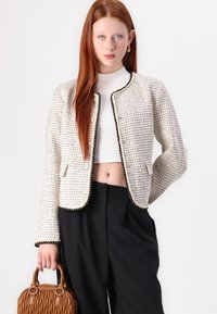 White tweed blazer with black trim, textured fabric, cropped design, two front pockets, paired with black trousers and a brown handbag.
