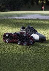 Black and white golf shoes on grass, one shoe showing metal spikes on the sole with a golf flag in the blurred background.
