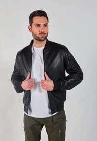 Veste bomber en cuir noir avec un col ajusté, des manches côtelées, une fermeture éclair à l'avant et des poches latérales. T-shirt blanc en dessous, mannequin en pose.