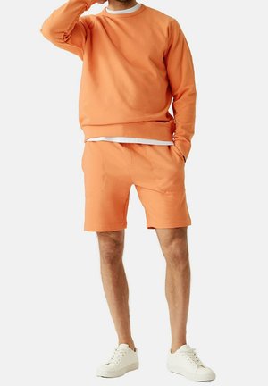 Hombre con sudadera naranja y pantalones cortos a juego, con zapatillas blancas, de pie con una mano en el bolsillo y la otra cerca de su rostro.