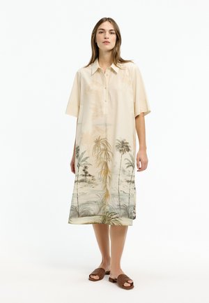 Woman wearing a beige knee-length dress with short sleeves and a landscape print of palm trees, paired with brown woven slide sandals.