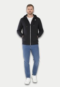 Haut à capuche zippé noir avec un logo blanc, deux poches avant et une capuche à cordon. Associé à un jean bleu clair et des baskets blanches.