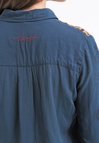 Vue arrière d'une personne portant une chemise bleue avec le logo "Desigual" brodé en rouge sous le col et un motif à motifs sur l'épaule.