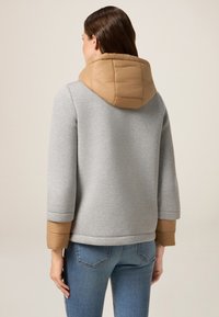 Felpa grigia con cappuccio e polsini in stile piumino di colore beige. Il tessuto appare liscio; il cappuccio ha una trama trapuntata. Indossata con jeans blu.