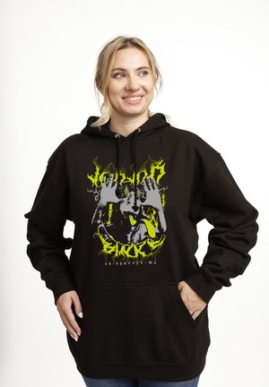 Uśmiechnięta kobieta w czarnej bluzie z kapturem z neonowo-żółtym i szarym nadrukiem przedstawiającym psa wystawiającego język i ręce opierające się o twarz.