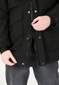 Mustat eristysjakut, joissa on vetoketju ja nepparit, kahdella etutaskulla ja sileällä pinnalla. Päällä tummanharmaat farkut.