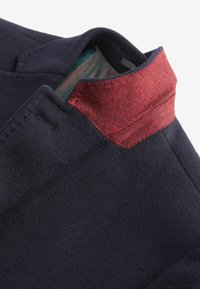 Abrigo de lana navy con cuello de fieltro rojo y forro con estampado en azul verdoso. Presenta una forma ajustada y detalles de costura limpios.