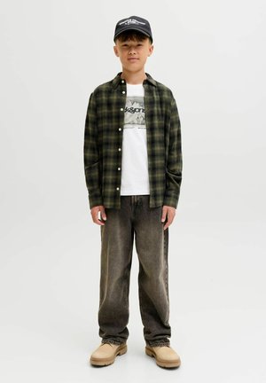 Jongen in een zwarte pet, groen geruit overhemd over een witte T-shirt met print, loszittende vervaagde zwarte spijkerbroek en beige boots, staande voor een witte achtergrond.