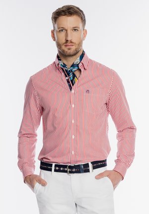 Mężczyzna ubrany w czerwoną i białą koszulę w paski na guziki, wzorzystą apaszkę na szyi, białe spodnie i czarny pasek, z rękami w kieszeniach.