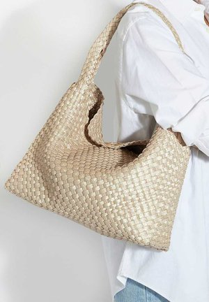 Beige woven shoulder bag with wide strap worn by person in white shirt and blue jeans, shown from side against plain background.