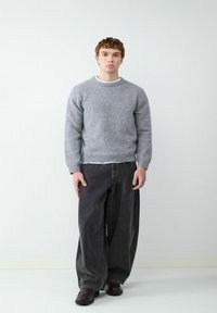 Grijze, gebreide trui met een ronde hals, gedragen over een wit shirt; gecombineerd met losse, donkere broeken en bruine schoenen. Over het geheel een gladde textuur.