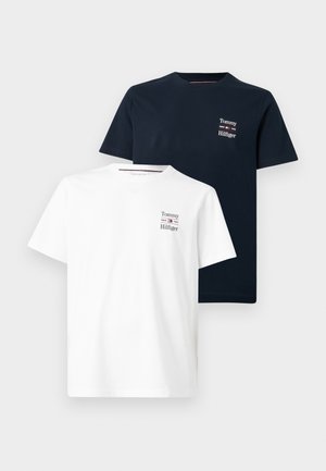 Mann mit brauner Mütze, schwarzem Tommy Hilfiger T-Shirt und Hose, beide mit weißem Logo, und weißen Turnschuhen, steht mit den Händen in den Taschen.