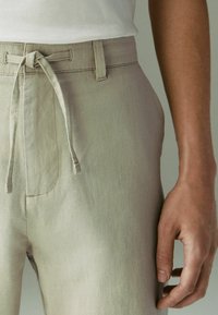 Pantalon en lin beige avec une taille à cordon noué, porté avec un haut blanc rentré à l'intérieur et une main détendue posée à côté de la hanche.