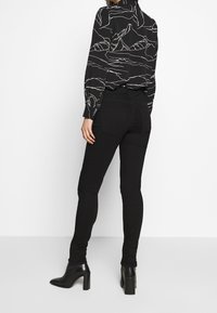 Svart blus med vitt bergsmönster, långa ärmar och hög krage, i kombination med svarta skinny jeans och högklackade ankelstövlar.