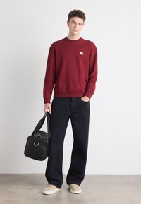 Felpa bordeaux con polsini a coste, pantaloni in denim scuro, sneakers beige e una borsa nera con accentuazioni in metallo argentato. Design semplice, vestibilità rilassata.