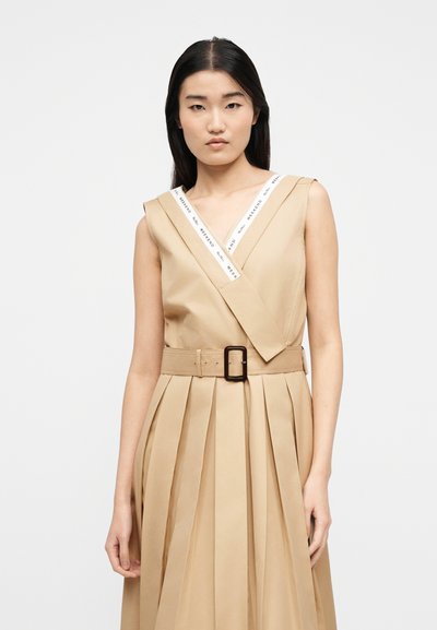 Νεαρή γυναίκα με μακριά σκούρα μαλλιά που φοράει αμάνικο μπεζ πλισέ φόρεμα με φαρδύ ζώνη και λευκή μπορντούρα με την επιγραφή "Weekend" στον γιακά.