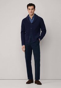 Tmavě modrý žebrovaný kardigan s šálovým límcem, zapínáním na knoflíky, dvěma předními kapsami, nošený přes modrou košili a kombinovaný s tmavými kalhotami a hnědými botami.