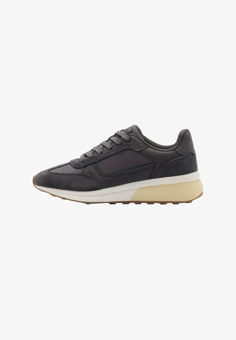 Grå atletisk sneaker med en kombination av släta och texturerade material, låg profil och en kontrasterande sula i vitt och tan.
