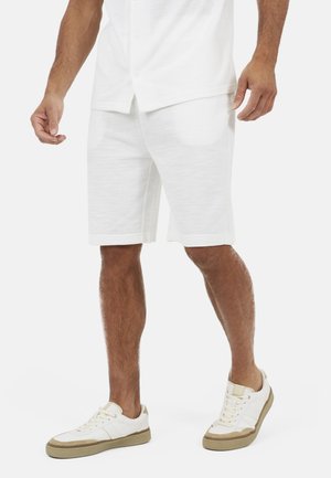 Homme portant un short décontracté blanc, une chemise blanche à manches courtes avec boutons, et des baskets blanches avec des semelles beige, debout sur un fond blanc.