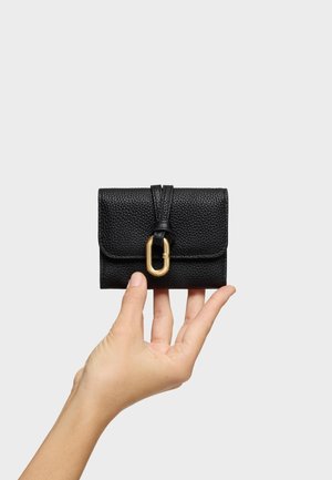 Mano che tiene un piccolo portafoglio in pelle nera texturizzata con una fibbia ovale dorata su uno sfondo grigio chiaro.