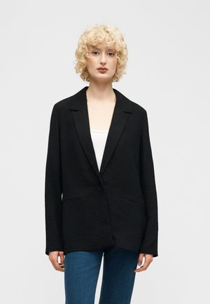 Vaaleatukkainen kiharatukkainen nainen, joka pukeutuu teksturoituun mustaan bleiseriin valkoisen paidan ja sinisten farkkujen päällä, seisoo tasaisen vaalean taustan edessä.