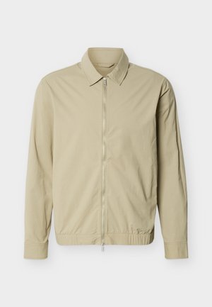 Bēša viegla jaka ar garām piedurknēm, apkakli un pilna garuma sudraba rāvējslēdzēju priekšpusē baltā fonā.