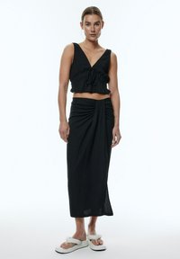 Schwarzes Cropped-Top mit V-Ausschnitt und Bindedetail, kombiniert mit einem langen schwarzen Rock mit gedrehtem Vorderdesign. Weiße Plateausandalen.