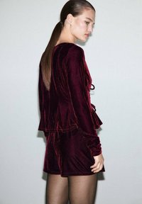 Woman with long brown ponytail wearing a fitted burgundy velvet long-sleeve top and matching shorts against a plain background.