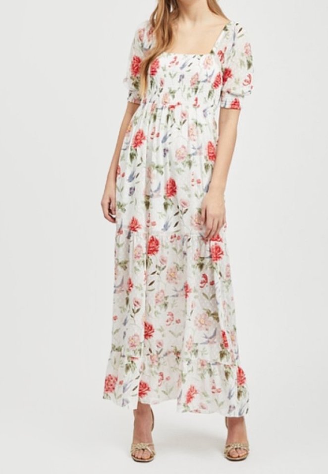 Femme portant une robe maxi blanche à fleurs avec manches bouffantes et décolleté carré, associée à des talons ouverts avec brides à la cheville.