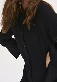 Abito nero a camicia con tessuto testurizzato, caratterizzato da un colletto e maniche ampie e fluide. Include una chiusura frontale con bottoni.