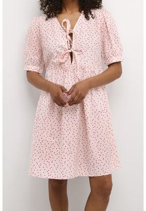 Kobieta ubrana w różową sukienkę do kolan z czerwonym nadrukiem serc, z krótkimi bufkowatymi rękawami i wiązaniami na dekolcie z przodu.