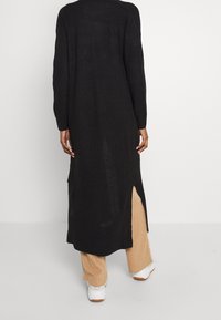 Schwarzer, langer Strickcardigan mit gerippter Textur, offenen Schlitzen an beiden Seiten und langen Ärmeln. Hat eine lockere Silhouette.