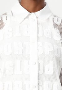 Λευκό πουκάμισο από διχτυωτό ύφασμα με γιακά που διαθέτει επαναλαμβανόμενο μοτίβο "SPORTS". Κουμπιά μπροστά, υφή υφάσματος και ανοιχτό πλέγμα.