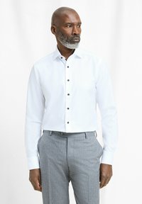Witte overhemd met lange mouwen en een klassiek kraag, voorzien van zwarte knopen, gecombineerd met grijze geblokte broek, gepresenteerd tegen een lichte achtergrond.