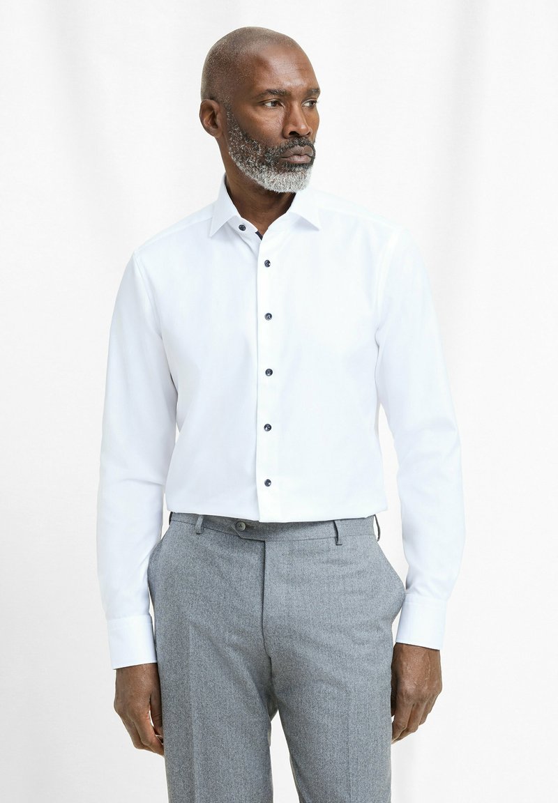 Witte overhemd met lange mouwen en een klassiek kraag, voorzien van zwarte knopen, gecombineerd met grijze geblokte broek, gepresenteerd tegen een lichte achtergrond.