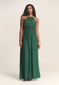 Dlouhé zelené šaty z hladké látky. Mají halterový výstřih, stažený pas se šňůrkami a široký maxi střih.